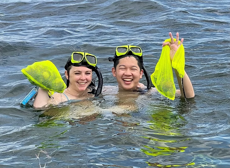 Two snorkelers in water holding yellow mesh bags and smiling.