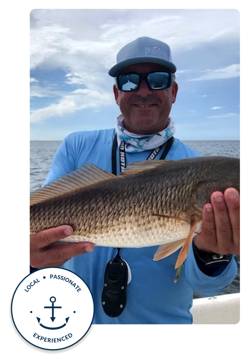 Man on a boat holding a large fish, wearing sunglasses and cap, with ocean in background.