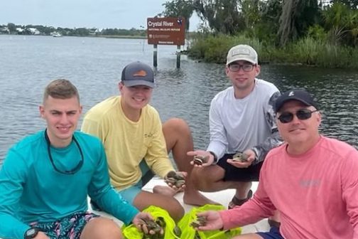 Four people on a boat holding bags of shells near the shore.